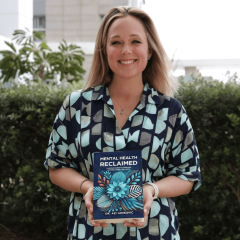 A photo of Dr Azi Jankovic standing in front of a bush holding her book, "Mental Health Reclaimed"